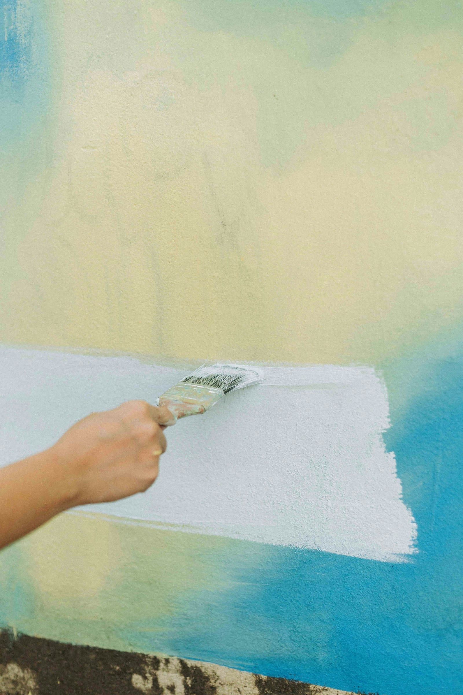 Painter's hand brushing white paint over a turquoise and yellow wall