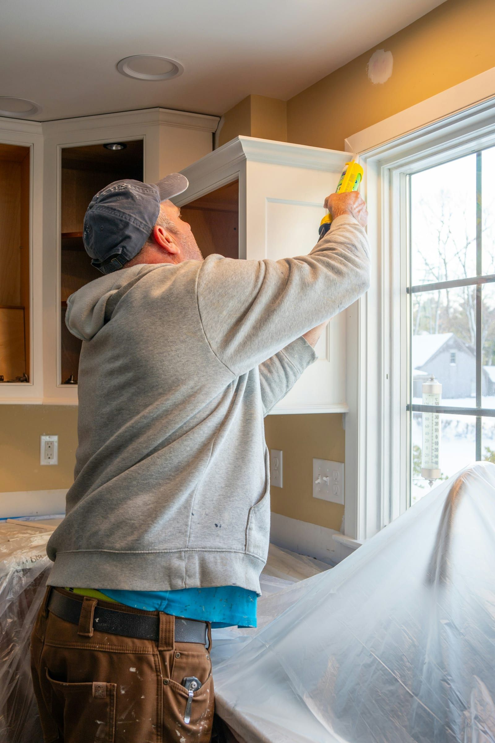 Painter caulking around a kitchen window frame indoors, back to the camera