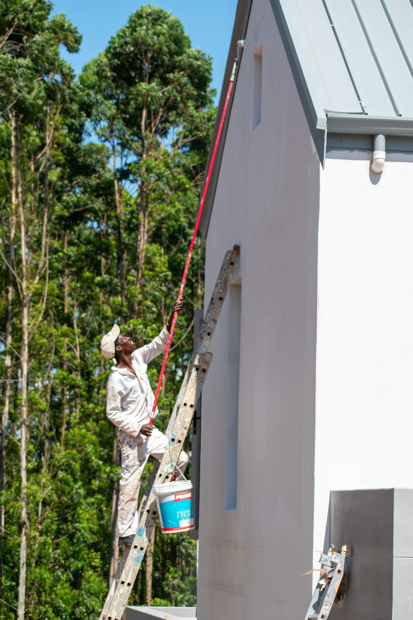 Painter on a ladder brushing a white stucco wall under a metal roof, trees behind