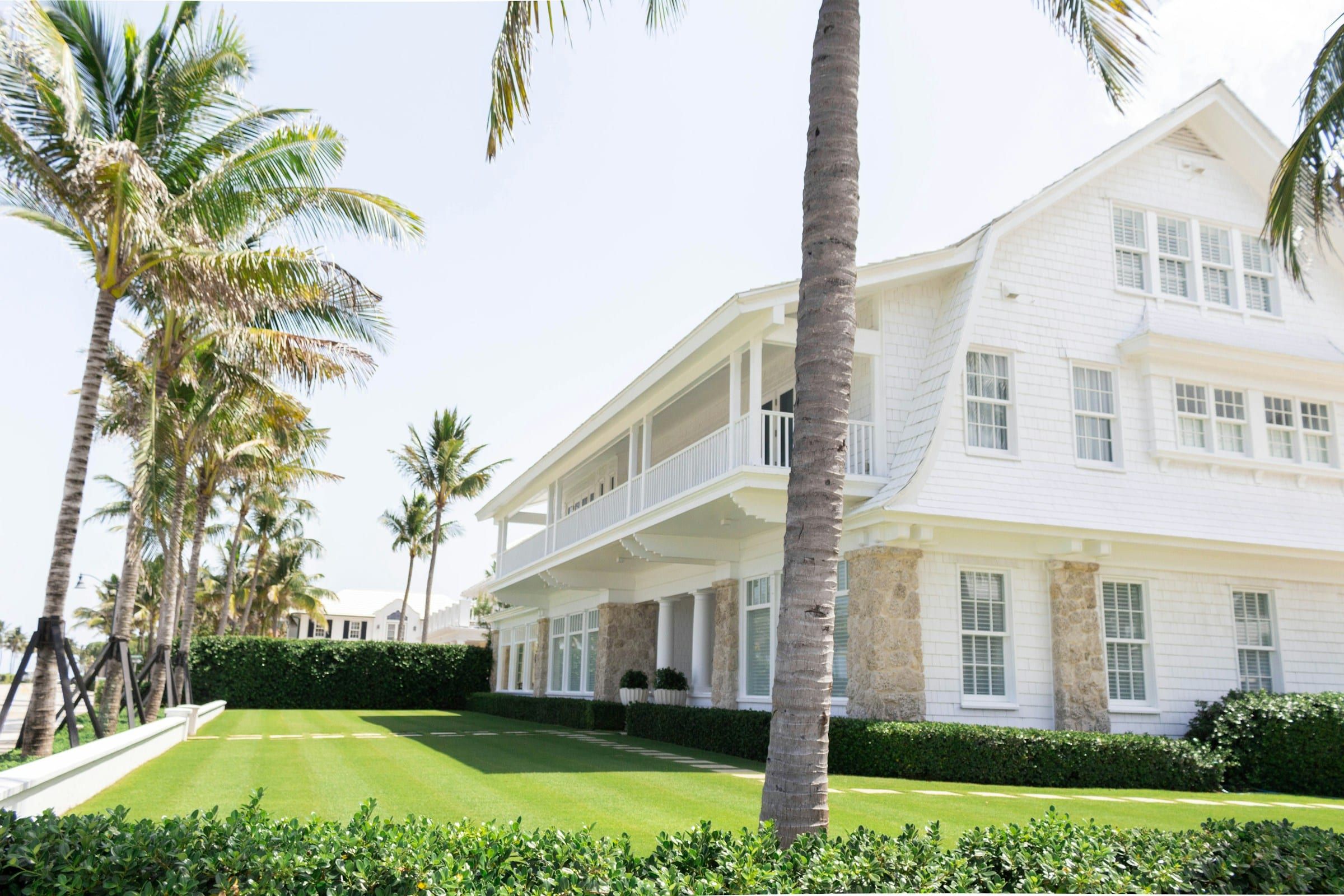 White wood-sided two-storey house with a lawn and palm trees under a clear sky